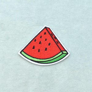 Può includere: Una toppa ricamata a forma di fetta di anguria. Il frutto triangolare presenta un interno rosso brillante con dettagli di semi neri, un bordo della buccia verde scuro e un contorno bianco, creando un design grafico chiaro e colorato.