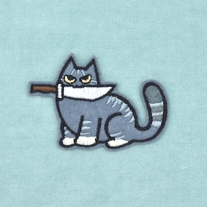 Op de afbeelding: Een geborduurde patch met een grijze cyperse kat met donkergrijze strepen, witte pootjes en gele ogen. De kat houdt een groot zilverkleurig mes met een bruin handvat in de bek, geplaatst op een effen lichtblauwe stoffen achtergrond.