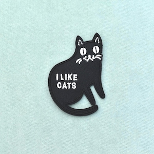 Könnte beinhalten: Ein schwarzer, bestickter Aufnäher in Form einer sitzenden Katze mit weißen Stickdetails. Die Katze hat große weiße Augen, Schnurrhaare und den Text I LIKE CATS in Weiß auf der Seite. Der Aufnäher hat einen sauber vernähten Randabschluss.