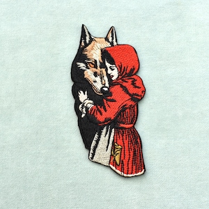 Peut inclure: Écusson brodé représentant un loup étreignant une figure vêtue d'une cape à capuche rouge. Le loup a une fourrure marron et noire, et la figure porte une robe rouge avec un tablier blanc. L'écusson est sur fond bleu clair.
