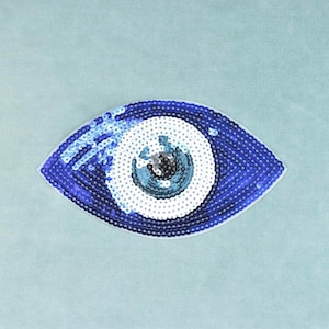 Puede incluir: Un parche con forma de ojo cubierto de lentejuelas que presenta un iris exterior azul oscuro, un anillo central ancho de color blanco y una pupila central de color azul claro y negro. El diseño está formado por lentejuelas reflectantes.