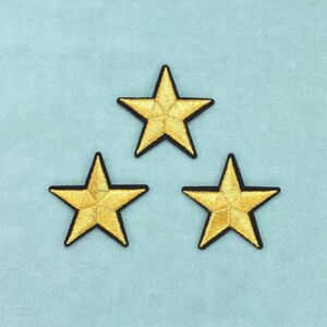 Può includere: Tre toppe ricamate a forma di stella con una finitura in filo dorato metallizzato e un distinto bordo nero. Ogni stella ha una forma a cinque punte con dettagli di cucitura visibili che si irradiano dal centro, disposte in formazione triangolare.