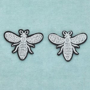 Puede incluir: Dos parches bordados de abeja con detalles en hilo metálico plateado sobre un borde de tela negra. Cada diseño de insecto muestra patrones de alas intrincados y un cuerpo segmentado, ofreciendo un aspecto de alto contraste para aplicar.