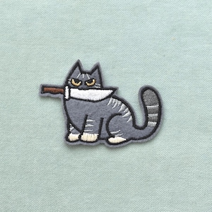 Op de afbeelding: Geborduurde patch met een grijze kat met een mes in de bek. De kat heeft gele ogen en witte accenten. De patch is zwart omlijnd en staat op een lichtblauwe achtergrond.