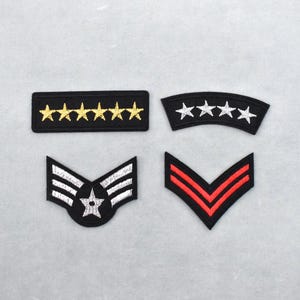 May include: Four embroidered military patches. The patches include a black rectangle with gold stars, a curved black patch with white stars, a black and white patch with a star, and a black patch with red stripes.