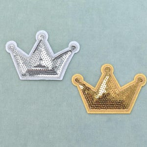 Könnte beinhalten: Zwei kronenförmige Aufnäher mit Paillettenoberfläche. Ein Aufnäher ist silberfarben mit weißem Rand, der andere ist goldfarben mit passendem goldenen Rand. Beide Kronen haben fünf Zacken mit kleinen kreisförmigen Akzenten an den Spitzen.
