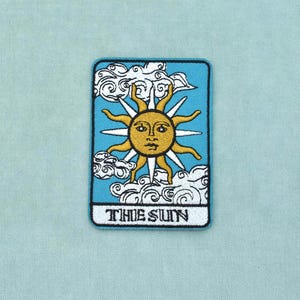 Peut inclure: Un écusson brodé rectangulaire représentant un soleil doré central avec un visage humain, entouré de nuages blancs sur un fond bleu clair. Le texte THE SUN est imprimé en lettres capitales noires sur une bannière rectangulaire blanche en bas.