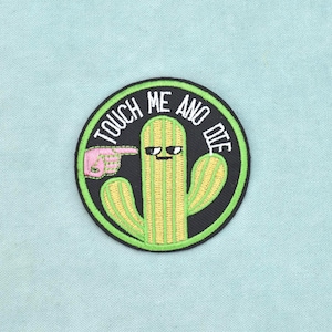 Puede incluir: Un parche bordado circular que presenta un cactus amarillo verdoso con una cara que lleva gafas de sol. Una mano rosa señala hacia el cactus. El texto "TOUCH ME AND DIE" está bordado en letras blancas a lo largo del borde superior sobre un fondo negro.
