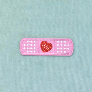 Op de afbeelding: Een rechthoekige roze pleister-patch met een centraal rood hartje met witte stippen. De patch heeft witte stikseldetails aan de linker- en rechterkant, die het uiterlijk van een standaard medische pleisterstrip nabootsen.
