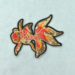 Peut inclure: Un écusson thermocollant en forme de poisson rouge, doté d'une broderie complexe dans des tons de rouge, orange et doré. Le motif est rehaussé de paillettes scintillantes sur tout le corps et les nageoires, le tout souligné par un bord noir.
