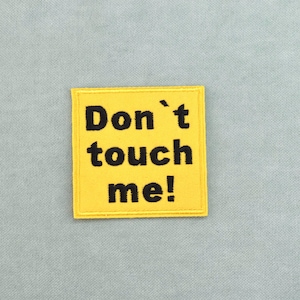 Peut inclure: Un écusson carré en tissu jaune avec une bordure cousue. L'écusson présente le texte brodé en noir "Don't touch me!" centré sur la surface. Le design est net et audacieux, avec le lettrage clairement visible sur le fond jaune.