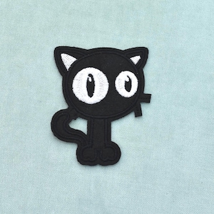 Op de afbeelding: Een zwarte geborduurde patch in de vorm van een cartoonkat met grote witte ronde ogen, puntige oren met witte details aan de binnenkant, een gekrulde staart en kleine voetjes. Het ontwerp heeft dikke zwarte stikselcontouren op een zwarte basis.