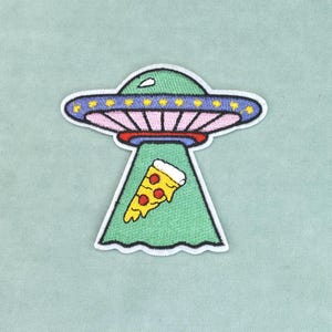 Op de afbeelding: Een geborduurde patch met een groene UFO met een paarse rand en gele lichten. De schotel zendt een groene tractorstraal uit die een punt pepperoni-pizza met gele kaas en rode toppings optilt tegen een lichtgroene achtergrond.