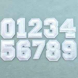May include: A set of ten white embroidered patches featuring the numbers zero through nine in a bold, block-style varsity font. Each patch has a clean, stitched border and a textured surface, arranged in two rows of five against a solid background.
