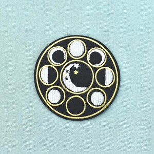 Peut inclure: Un écusson brodé circulaire avec un fond noir et une bordure dorée. Le centre présente un croissant de lune blanc avec de petites étoiles. Autour du centre se trouvent sept petites icônes circulaires représentant diverses phases lunaires en blanc.