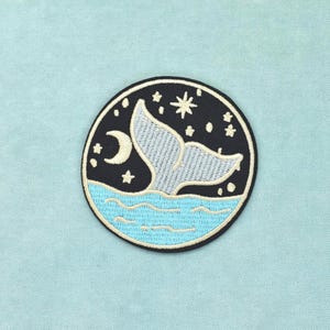 Può includere: Una toppa ricamata circolare raffigurante una coda di balena azzurra che emerge da onde marine azzurre su un cielo notturno nero. La scena include una luna crescente bianca e diverse stelle bianche di varie dimensioni nel bordo.
