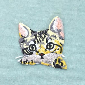 Pode incluir: Um emblema bordado apresentando a cabeça e as patas dianteiras de um gatinho malhado. O gatinho tem padrões de pelo amarelo, castanho e preto, olhos grandes e escuros, orelhas com tons rosados e um nariz pequeno e escuro com bigodes brancos.