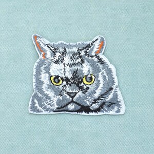 Peut inclure: Un écusson brodé représentant la tête d'un chat gris avec une texture de fourrure détaillée, des yeux jaune vif et de petites touches orange à l'intérieur des oreilles. Le chat a une expression sérieuse avec des ombres gris foncé.