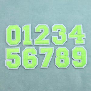 Op de afbeelding: Een set van tien opstrijkbare patches met de cijfers nul tot en met negen in een heldere neon-groene kleur. Elk cijfer heeft een dik, blokvormig varsity-lettertypeontwerp met een lichtgrijze rand die het levendige groene stoffen oppervlak omlijnt.