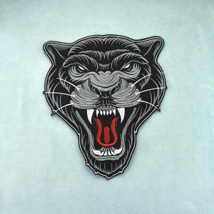 May include: An embroidered patch featuring the head of a black panther with its mouth open, revealing white teeth and a red tongue. The design includes detailed grey stitching for the fur texture, long white whiskers, and a black border.