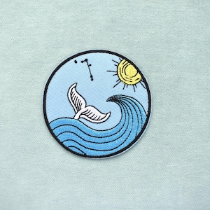 May include: A round, embroidered patch featuring a whale tail emerging from blue waves under a yellow sun. The design includes a constellation and is set against a light blue background with a black border.