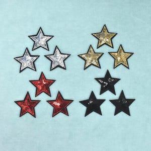 Peut inclure: Un ensemble de douze écussons en forme d'étoile disposés en quatre groupes de trois. Le lot comprend des étoiles argentées, dorées, rouges et noires, chacune présentant une surface à paillettes avec un contour noir distinct sur les bords.