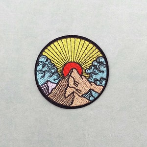 May include: Embroidered patch depicting a mountain range with a red sun rising behind it. The mountain is brown and the sun is red. The background is blue and yellow.