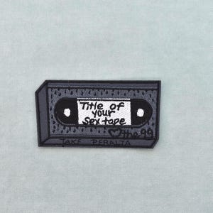 Könnte beinhalten: Gestickter Aufnäher in Form einer grauen und schwarzen Kassette. Der Aufnäher trägt den weißen Text "Title of your Sextape". Der Name "Jake Peralta" steht darunter.