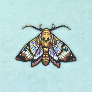 Puede incluir: Un parche bordado que representa una polilla con una marca similar a una calavera en el tórax. Las alas tienen tonos marrones, beige y violeta claro con contornos y detalles negros. Presenta dos antenas finas y curvadas sobre la cabeza.