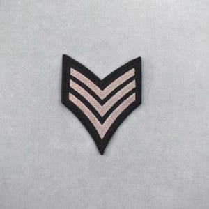 Peut inclure: Un écusson en forme de chevron noir et beige avec trois chevrons. Le patch est en forme de V, avec une bordure noire et des rayures beiges. Il est probablement destiné à un uniforme militaire ou de service.