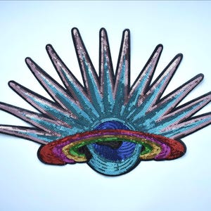 May include: A large, sequined patch featuring a stylized sunburst design. The sun has blue and pink rays, a blue centre, and a rainbow arching below. The patch is outlined in black.