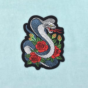 Peut inclure: Un écusson brodé représentant un cobra enroulé aux écailles bleues et blanches, avec un œil jaune et la gueule ouverte montrant une langue rouge. Le serpent est entouré de trois roses rouges à feuilles vertes et de petits boutons floraux verts.