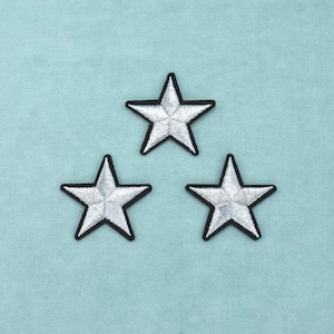 May include: Three embroidered star patches featuring a metallic silver thread finish and a distinct black border outline. Each star has a five-pointed shape with a textured, stitched surface that creates a subtle geometric pattern across the center.