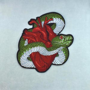 Pode incluir: Um emblema bordado apresentando um coração humano anatômico detalhado em tons de vermelho profundo, firmemente enrolado por uma cobra verde com escamas ventrais brancas e um olho vermelho. O design é contornado por uma borda preta espessa.
