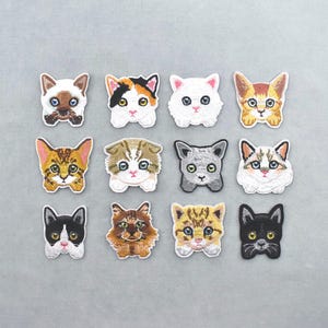Op de afbeelding: Een verzameling van twaalf geborduurde kattenkopjes in verschillende kleuren en patronen. De patches tonen gedetailleerde gezichtsuitdrukkingen en zijn gerangschikt op een lichtgrijze achtergrond. Elke patch is ongeveer 3,8 cm breed.