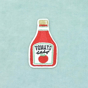 May include: An embroidered patch shaped like a bottle of tomato ketchup. The patch features a red bottle with a gold cap, a white label with the black text TOMATO ketchup, and a small red tomato illustration centred on the white background.
