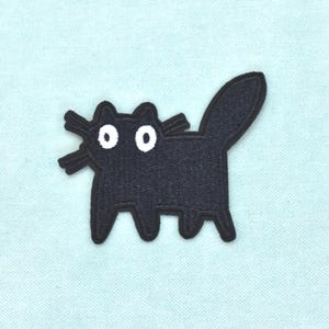Könnte beinhalten: Ein gestickter Aufbügler in Form einer stilisierten schwarzen Katze mit einfachem Umriss, erhobenem Schwanz und zwei großen weißen kreisförmigen Augen mit schwarzen Pupillen. Die Katze hat drei Schnurrhaare auf jeder Seite und vier kurze Beine.