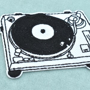 May include: A rectangular embroidered patch featuring a black and white line art design of a turntable. The patch displays a central black vinyl record on a white base with detailed outlines of the tonearm, platter, and control knobs.