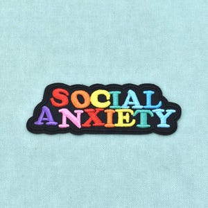 Può includere: Una toppa ricamata con sfondo nero che presenta il testo SOCIAL ANXIETY in lettere multicolori. La parola SOCIAL si trova sulla riga superiore in rosso, giallo e blu, mentre ANXIETY si trova sulla riga inferiore in viola, rosa, rosso, arancione, giallo e blu.