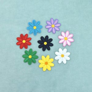 May include: A collection of eight embroidered flower patches arranged in a circular pattern. Each patch features a distinct color including blue, purple, pink, white, yellow, green, red, and black, all with a contrasting yellow or orange center.