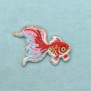 Peut inclure: Un écusson brodé en forme de poisson rouge avec un corps rouge, des écailles dorées et une queue fluide aux dégradés de rose, rouge et bleu clair. Le poisson est entièrement bordé d'un fil métallique doré scintillant.