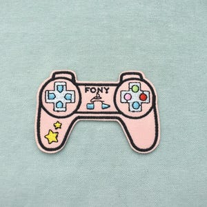 Pode incluir: Um patch bordado rosa em forma de controle de videogame. O patch tem contornos pretos e apresenta a palavra "FONY" acima do centro. Os botões do controle são de várias cores e há estrelas amarelas no lado esquerdo.