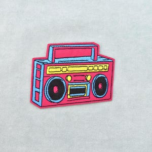 Könnte beinhalten: Ein farbenfroher Aufnäher mit einem Retro-Boombox-Design. Der Aufnäher hat einen pinkfarbenen Körper mit blauen und gelben Akzenten und schwarzen Lautsprecherdetails. Die Boombox hat einen Griff und Lautsprechergitter. Ideal, um Kleidung oder Accessoires einen Hauch von Vintage zu verleihen.