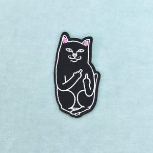 Peut inclure: Un écusson brodé représentant un chat noir aux contours blancs, avec l'intérieur des oreilles et le nez roses. Le chat est représenté en position assise, les deux pattes avant levées, chacune montrant un doigt d'honneur provocateur.