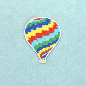 Könnte beinhalten: Ein gestickter Aufnäher in Form eines Heißluftballons. Der Ballon zeigt ein sich wiederholendes Muster aus diagonalen, zickzackförmigen Streifen in leuchtenden Farben wie Rot, Gelb, Hellblau und Grün, mit einem kleinen gelben Korb an der Basis.
