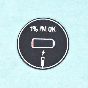 Può includere: Una toppa circolare ricamata nera con un bordo bianco. Il design presenta un'icona di batteria scarica con un piccolo indicatore rosso, un simbolo di cavo di ricarica e il testo bianco 1% I'M OK stampato sopra il grafico della batteria.