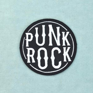 Op de afbeelding: Een ronde, zwart geborduurde patch met de witte tekst PUNK ROCK in een gestileerd lettertype. Het ontwerp is omsloten door een dunne witte cirkelvormige rand, wat een grafisch element met hoog contrast vormt voor toepassing op diverse stoffen.