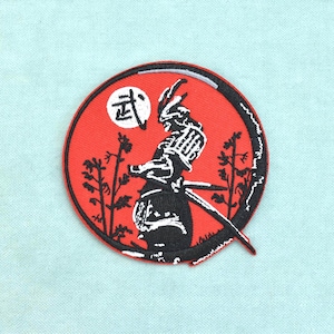 Könnte beinhalten: Ein kreisförmiger bestickter Aufnäher mit rotem Hintergrund und der schwarzen Silhouette eines Samurai, der zwischen Bäumen steht. Ein weißer Kreis mit dem schwarzen japanischen Kanji-Zeichen für Kampfkunst befindet sich oben links.