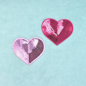 Op de afbeelding: Twee hartvormige patches bedekt met lovertjes. Het ene hart is fel, diep magentarood, terwijl het andere hart een gespleten ontwerp heeft met de ene helft in lichtroze en de andere helft in een donkerdere, metallic roze tint.