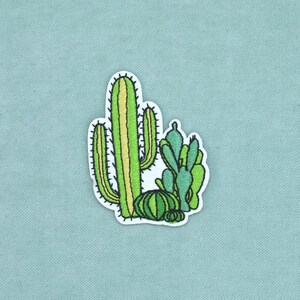 May include: An embroidered patch featuring a cluster of various cacti in shades of lime, forest, and emerald green. The design includes a tall columnar cactus, a round barrel cactus, and several smaller prickly pear pads with dark outlines.